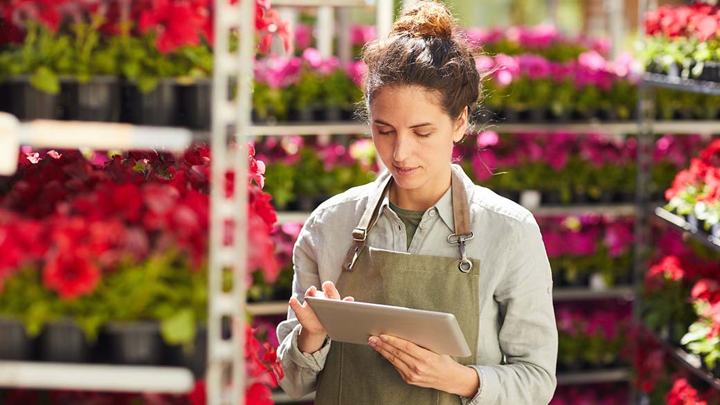 Waist up portrait of young woman using digital tablet surrounded by beautiful flowers while enjoying work in plantation outdoors
